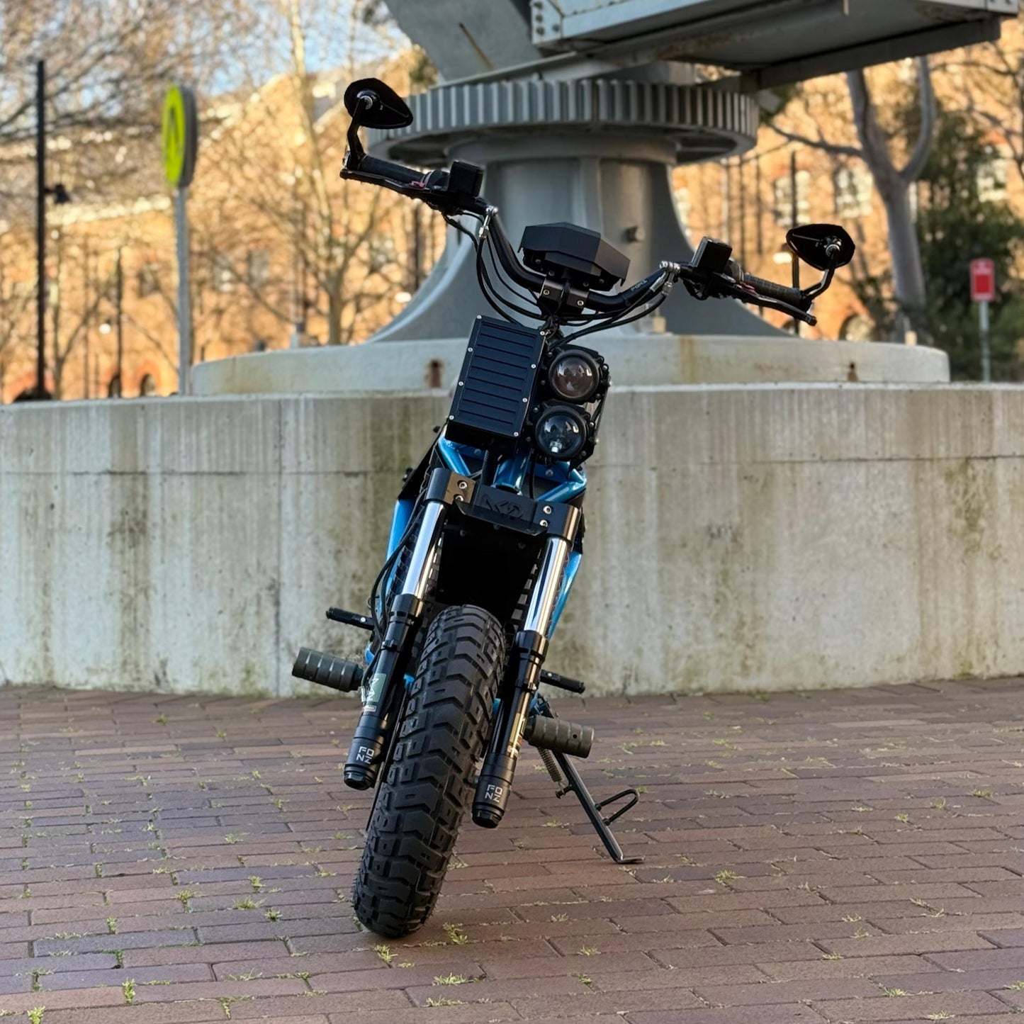 FONZ NKDs Electric Motorcycle on a stand in front of a large metal sculpture with trees and a building in the background.